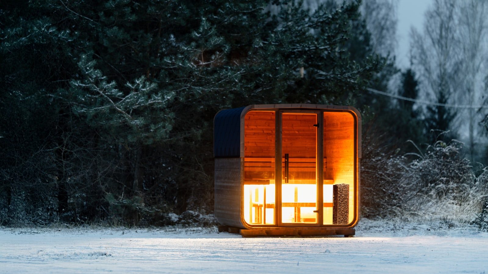 Luxury glass-front outdoor estate sauna glowing warm at dusk in a snowy forest clearing