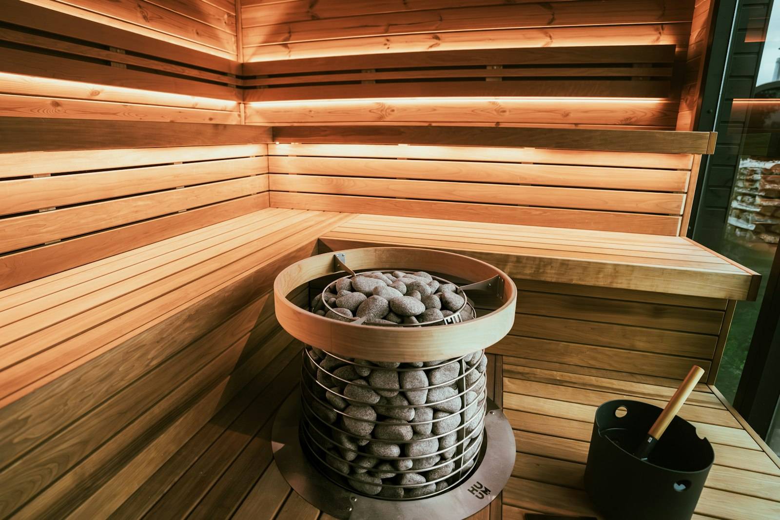 Modern cylindrical sauna heater filled with stones beside cedar benches and a water bucket